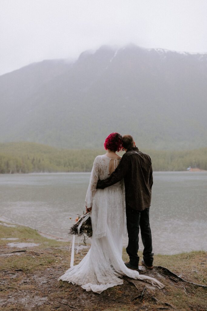 A couple hugs while overlooking scenic mountain views of Mirror Lake, one of the best places to elope in Alaska.