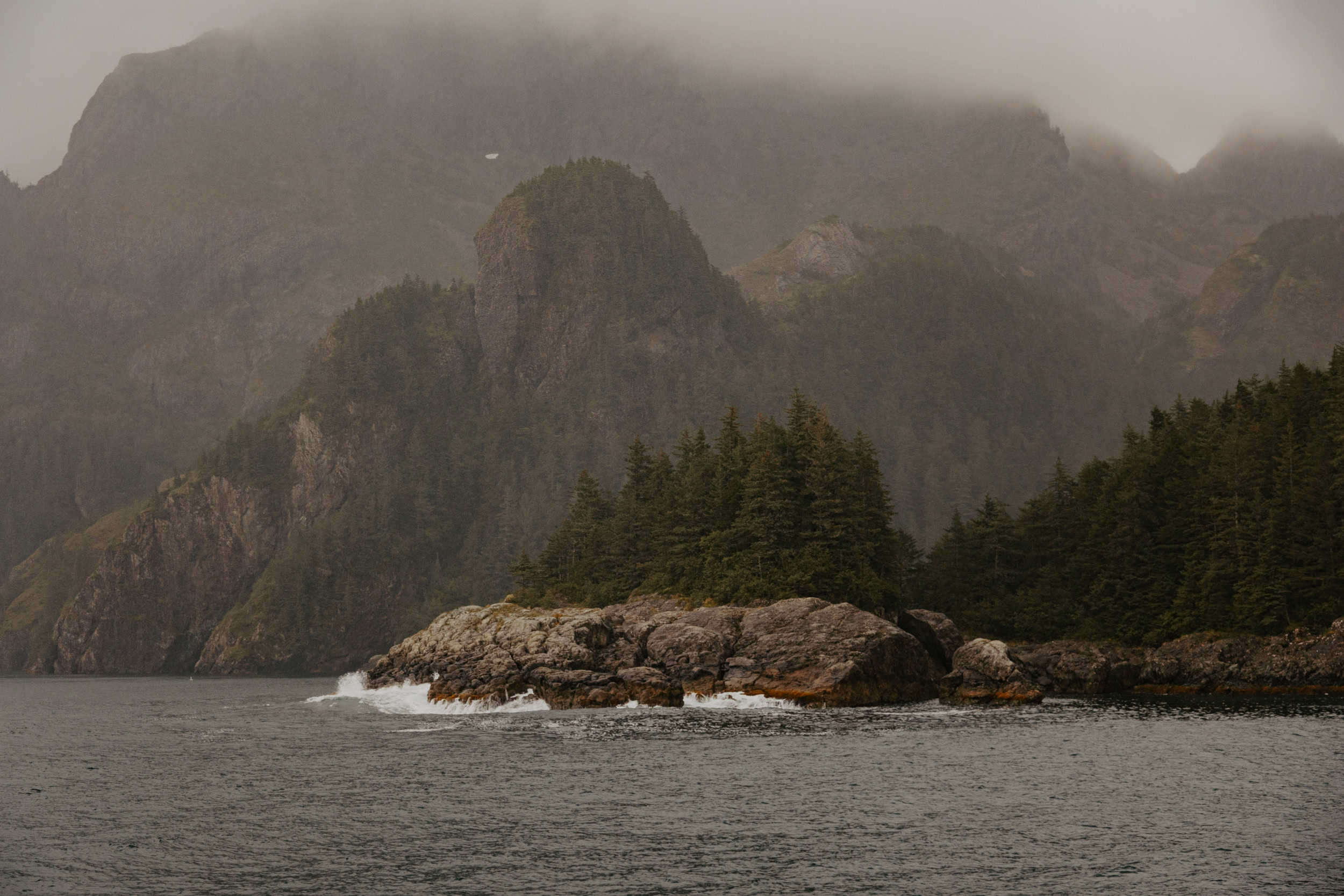 Waves crash into a forested cliffside with tall mountains in the background in Kenai Fjords National Park.