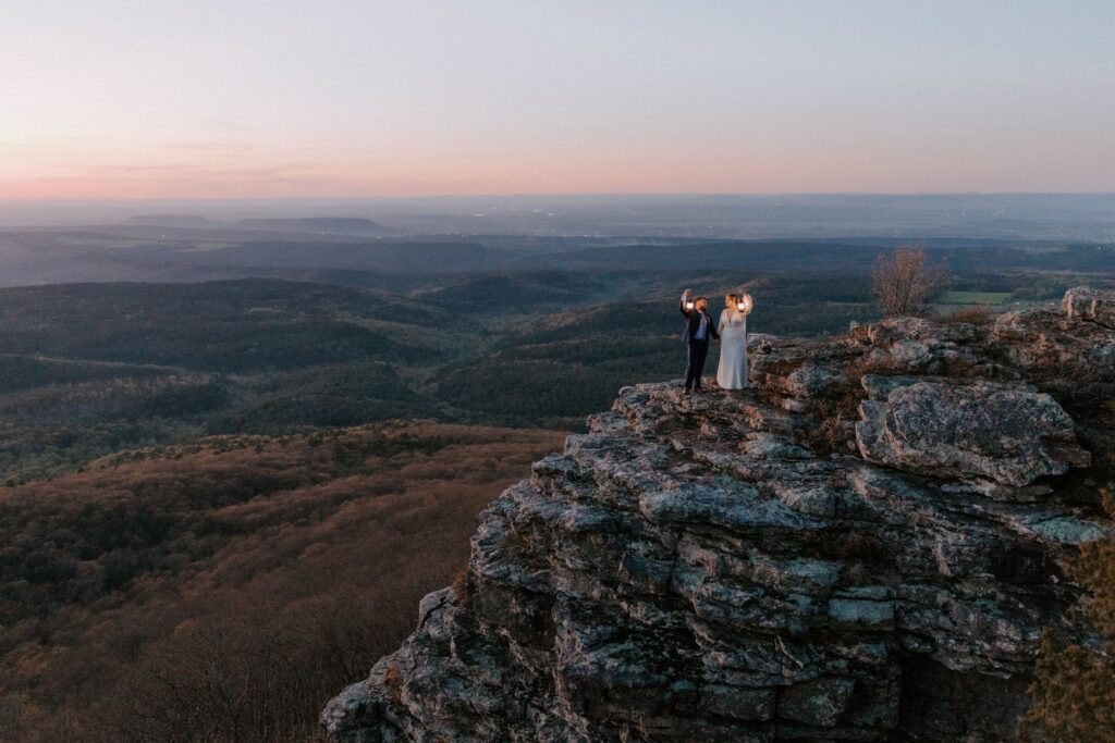 A couple stands on a cliffside at Mount Magazine during sunset while holding lanterns to see their path.
