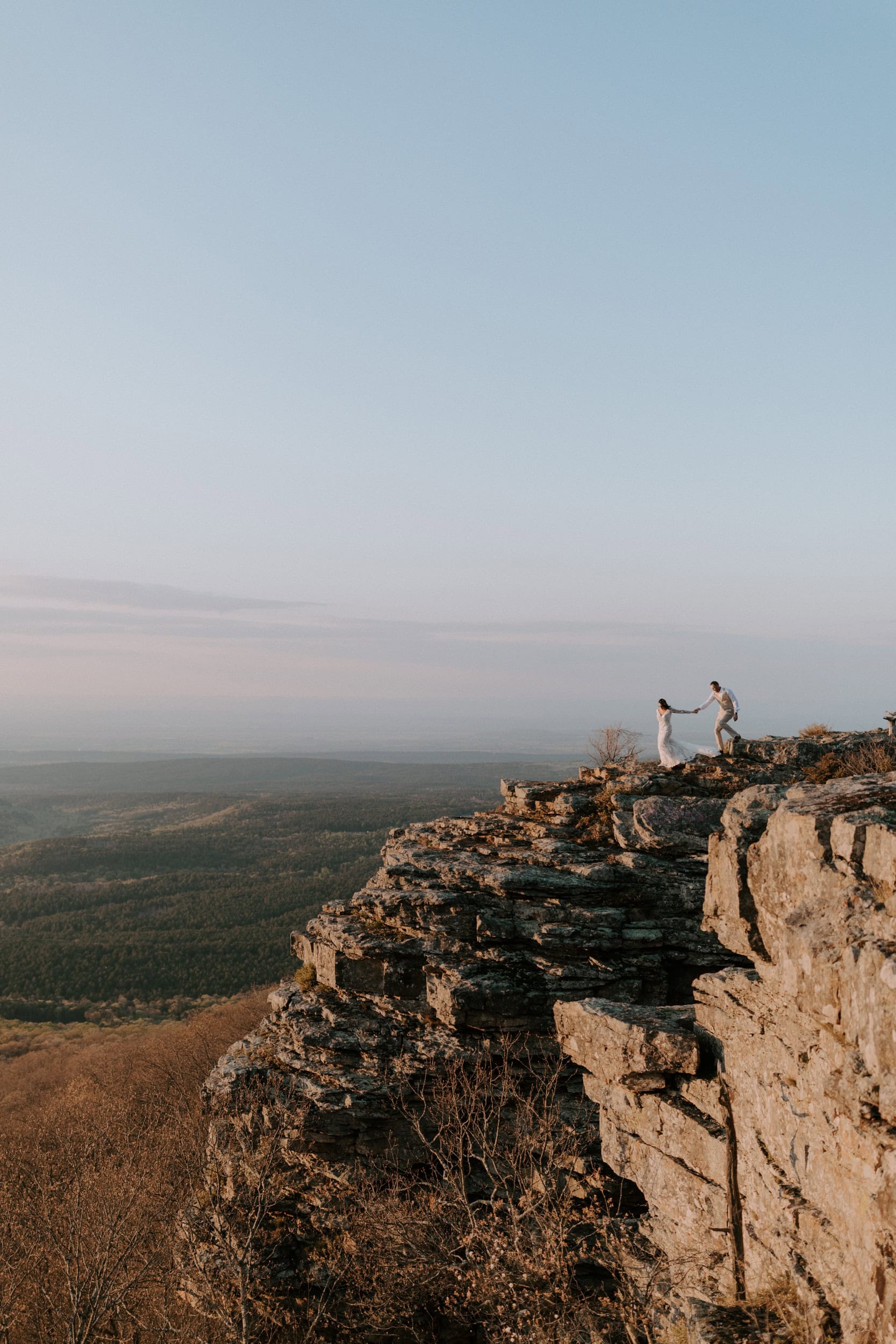 A couple in wedding attire is seen at a distance walking down a cliffside during their Arkansas Elopement at Mount Magazine State Park on Cameron Bluff.