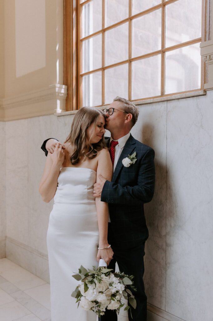 A couple stands under a large window at Pulaski County Courthouse in Little Rock, Arkansas. The groom is kissing his bride on the temple after their elopement ceremony.