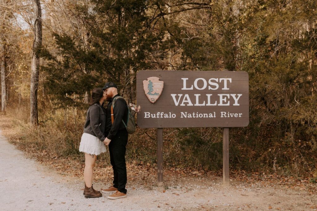 A couple kisses by the Lost Valley trailhead sign in the Buffalo National River area before their Lost Valley elopement adventure.