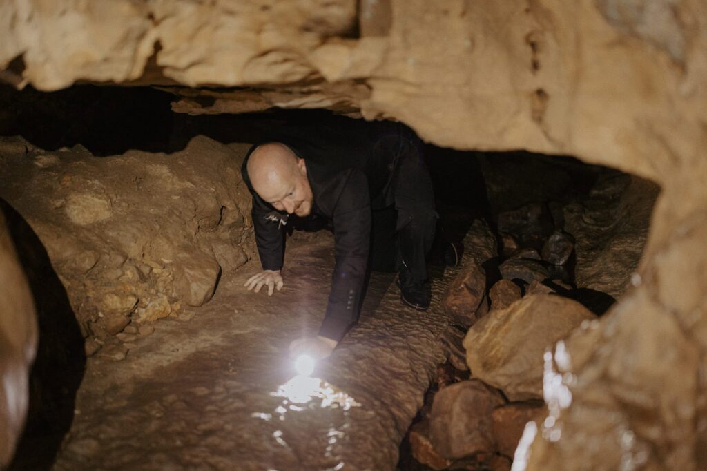 A groom crawls on all fours with a flashlight while exploring Lost Valley Trail in Arkansas.