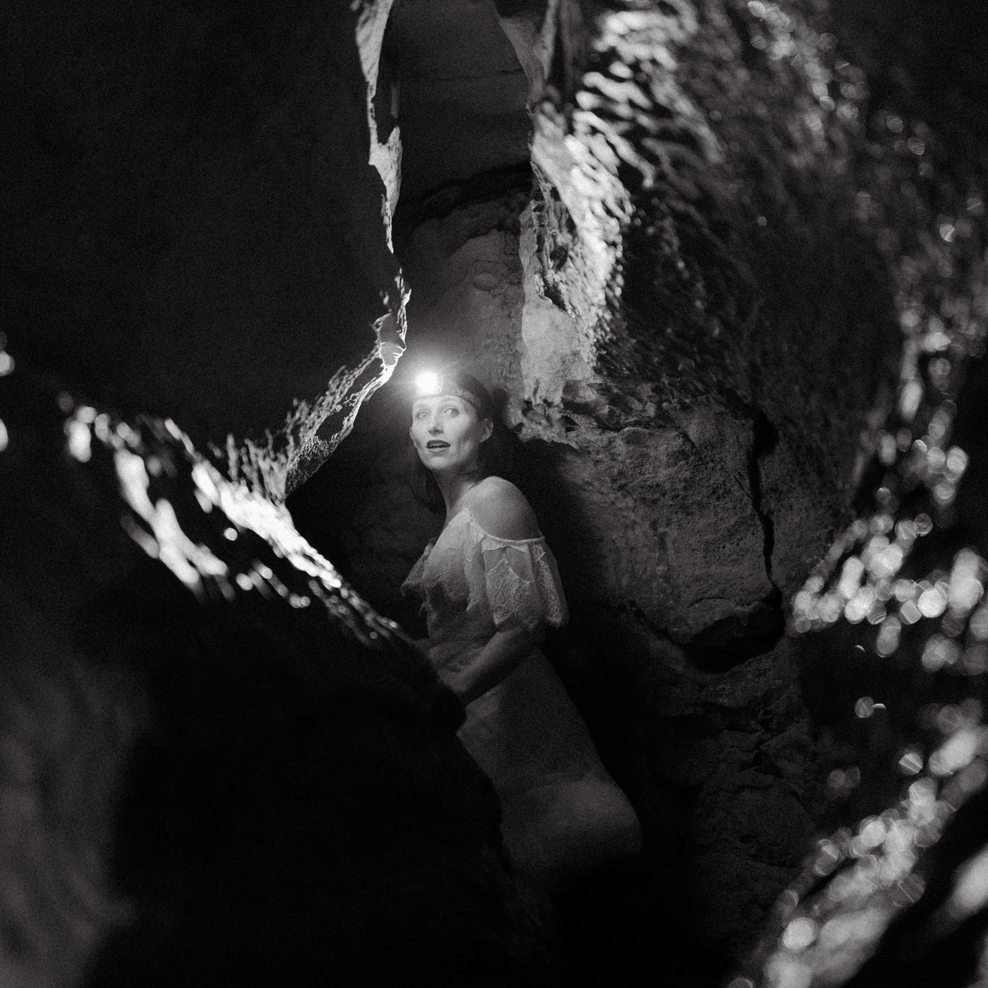 A bride walking through Eden Falls Cave at Lost Valley trail during her Arkansas elopement looks back at the camera and lights up the walls with her headlamp.