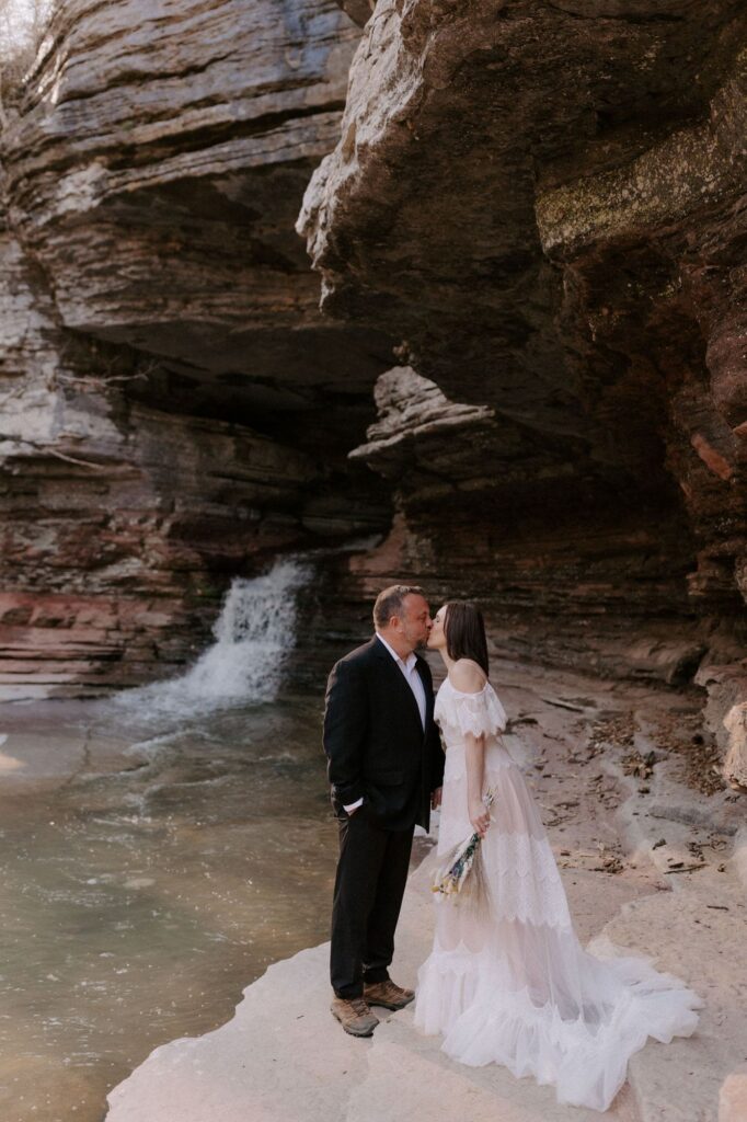 A couple kisses below the Natural Bridge waterfall at Lost Valley in Ponca, Arkansas while exploring for their elopement.