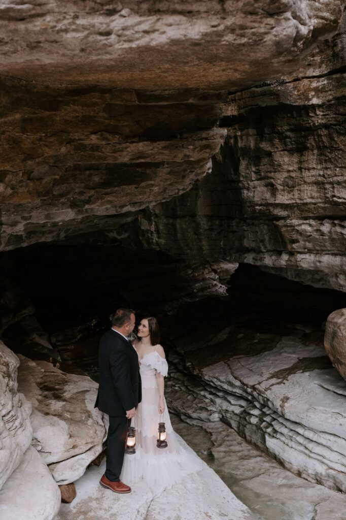 A couple stands outside the entrance of the Natural Bridge at Lost Valley Trail with lanterns while exploring for their elopement in Arkansas.