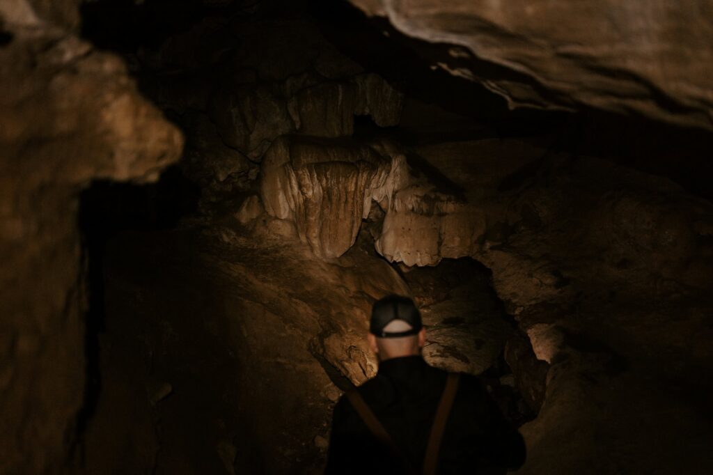 a groom peaks down into a cave with a flashlight at Lost Valley Trail while exploring for his adventure elopement.