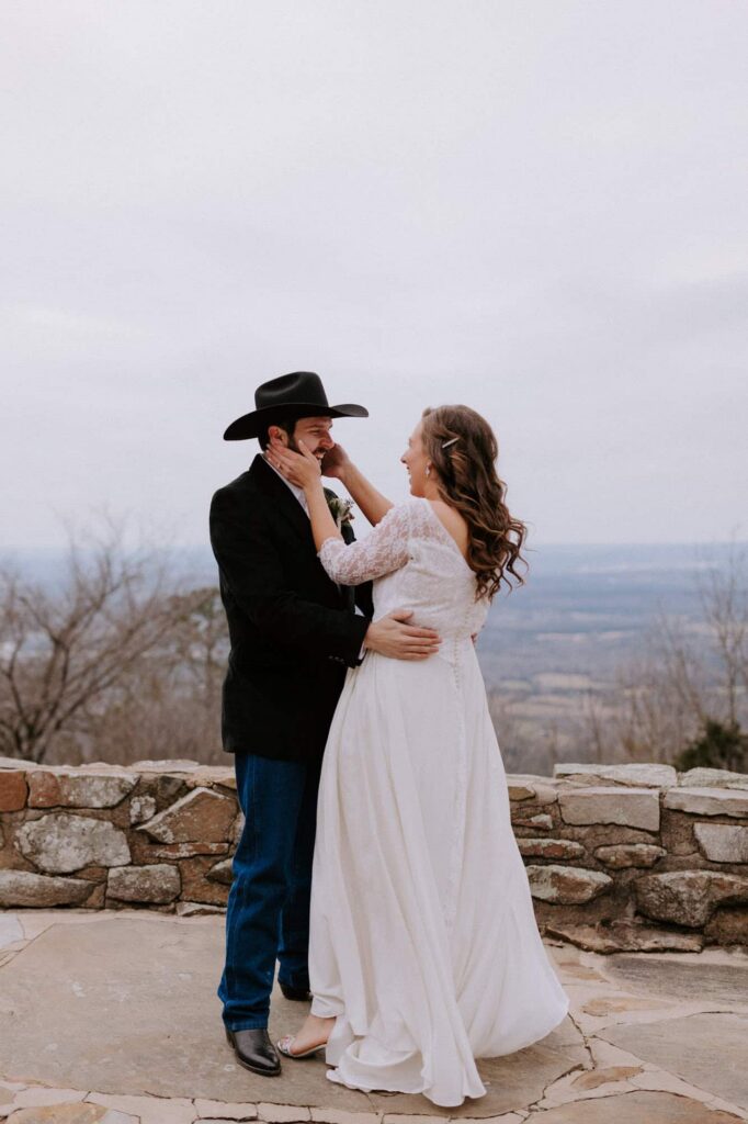 A groom laughs in excitement while his bride grabs his face with both hands during their first look at an overlook at Mt. Nebo State Park.
