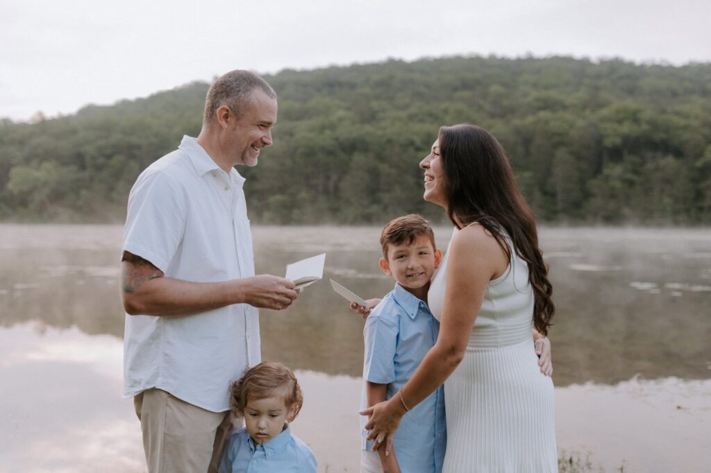 A husbnad and wife stand beside a lake with their two children, laughing as they read their vows to each other during their vow renewal in Arkansas.
