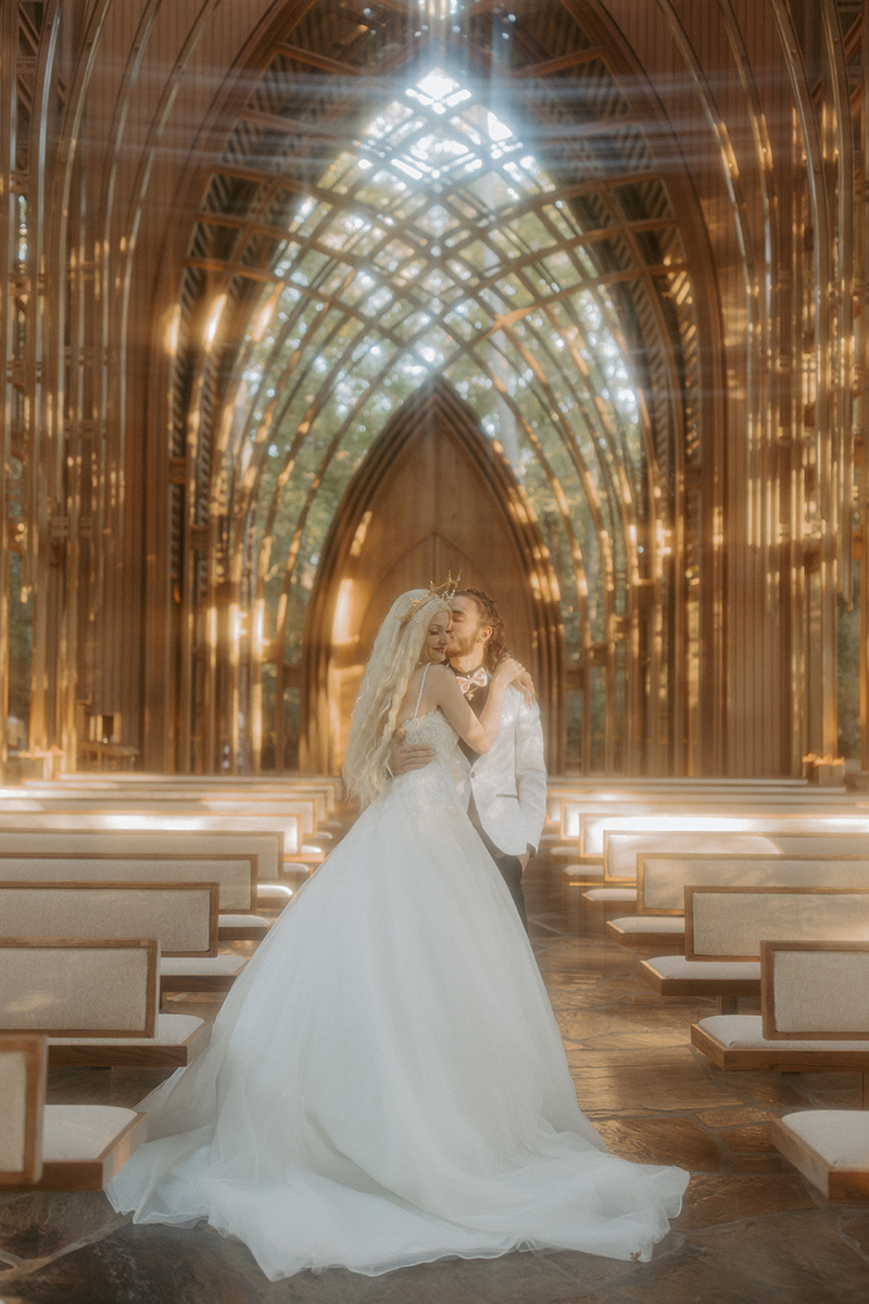 A couple in wedding attire are hugging each other in the middle of the aisle of Cooper Chapel, a glass chapel located in Bella vista, Arkansas. Sunlight is pouring in through the glass.