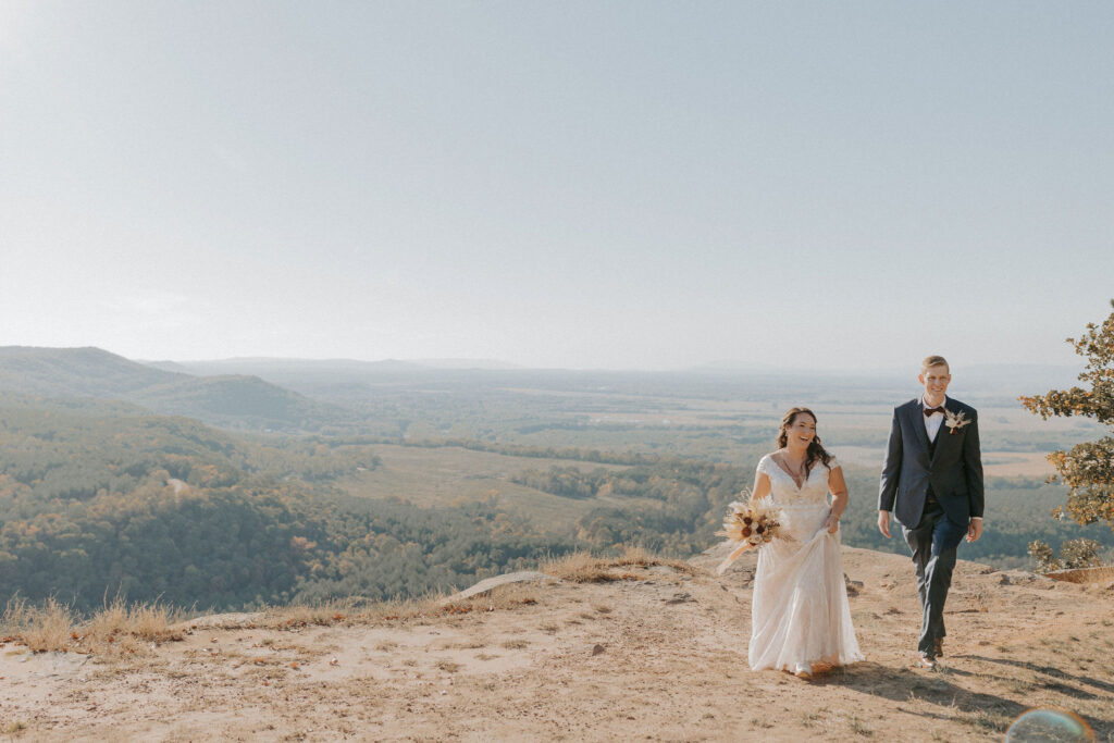 A couple in wedding attire walk towards the camera at CCC Overlook at Petit Jean State Park in Arkansas. The bride is in a white floor-length dress holding a dried floral arrangement and the groom is in a navy blue suit.