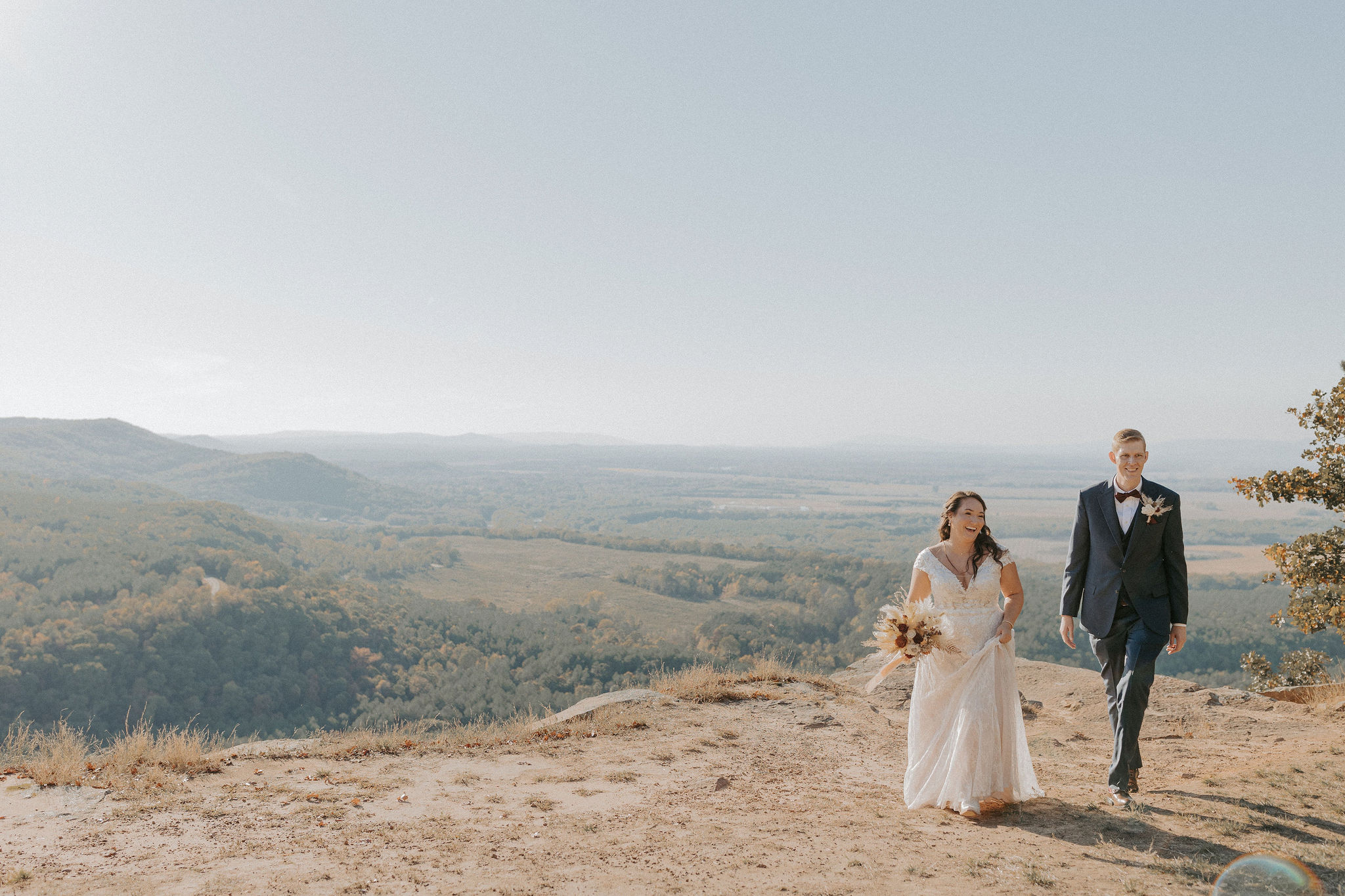 A couple in wedding attire walk towards the camera at CCC Overlook at Petit Jean State Park in Arkansas. The bride is in a white floor-length dress holding a dried floral arrangement and the groom is in a navy blue suit.