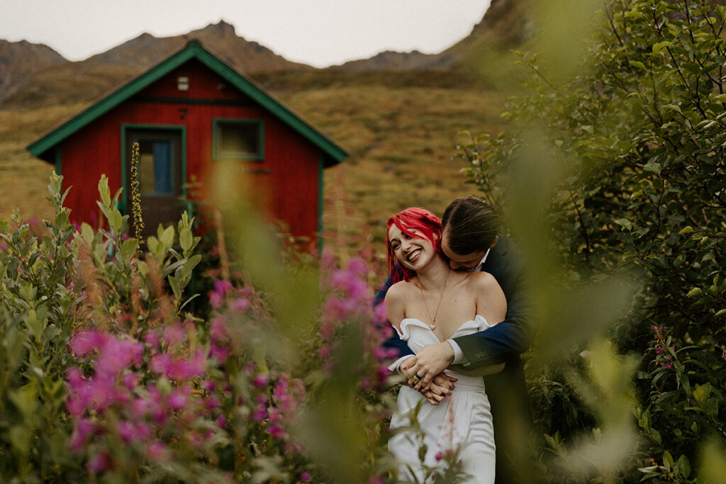 A couple in wedding attire are embracing in the middle of fireweed at Hatcher Pass in Alaska for their elopement.